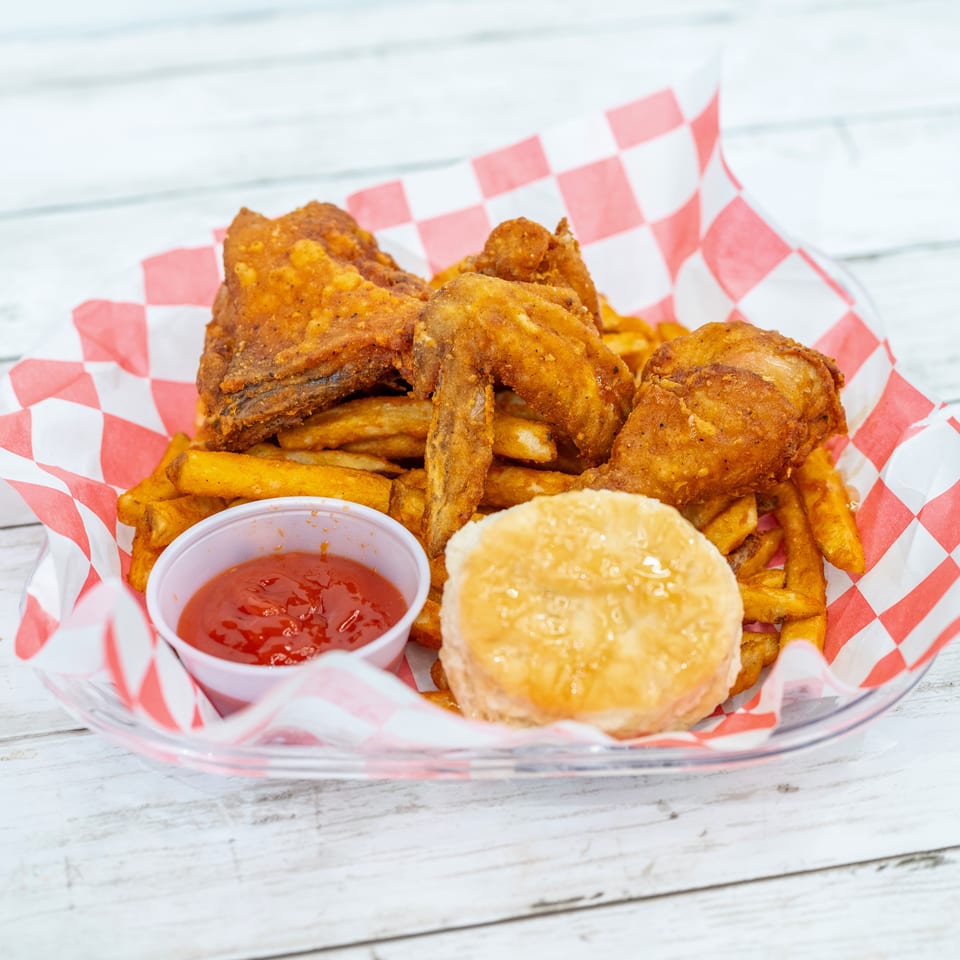 Basket of crispy fried chicken and French fries with ketchup and a biscuit on red-and-white checkered paper.