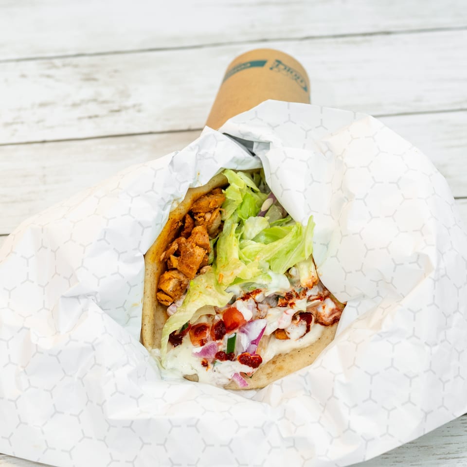Open chicken gyro wrap with lettuce, onions, tomatoes, and creamy sauce in patterned paper on a white wooden table.