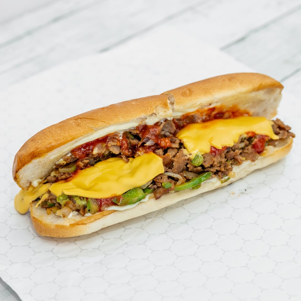 Philly cheesesteak sandwich with sliced beef, melted cheese, peppers, onions, tomatoes, and sauce on a toasted roll.