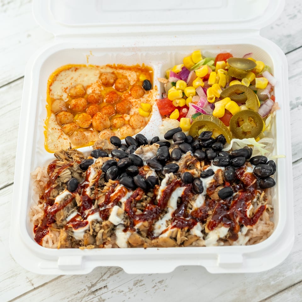 Open takeout container filled with rice, shredded meat, black beans, hummus, corn salad, jalapeños, and drizzled sauces.