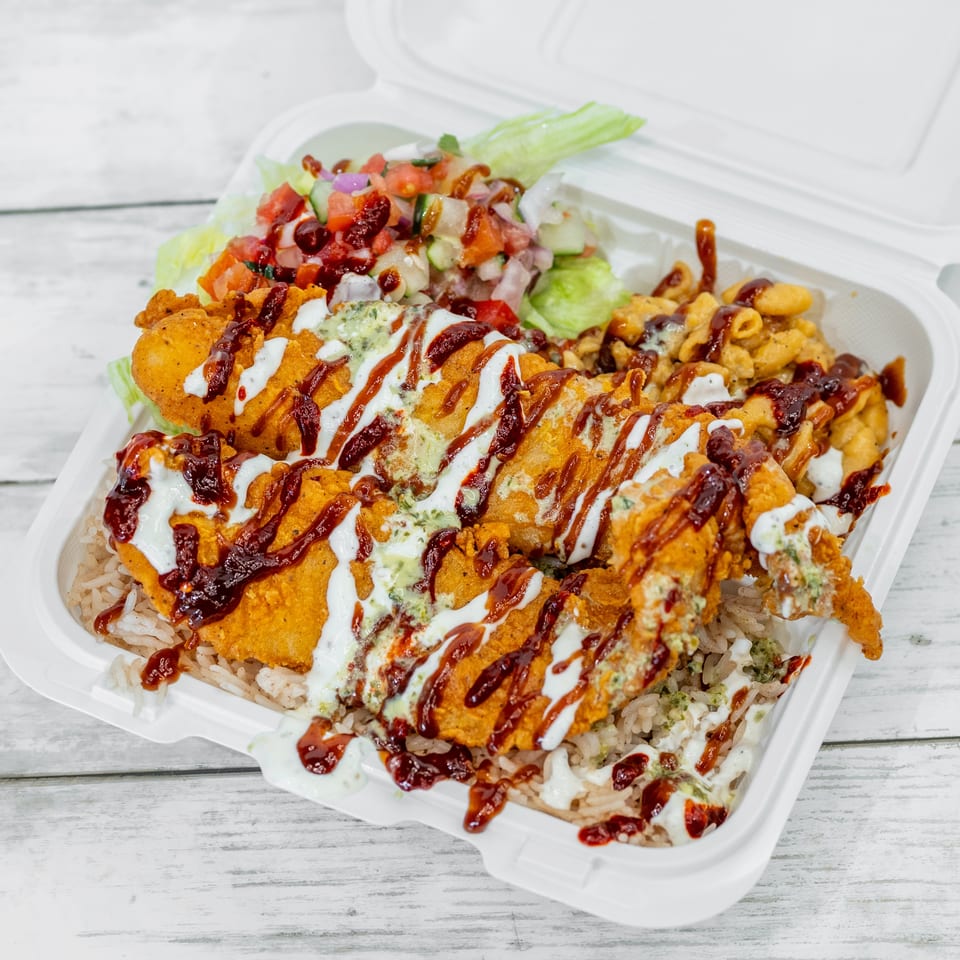 Takeout container filled with crispy fried chicken, rice, salad, macaroni, and drizzled white and red sauces.