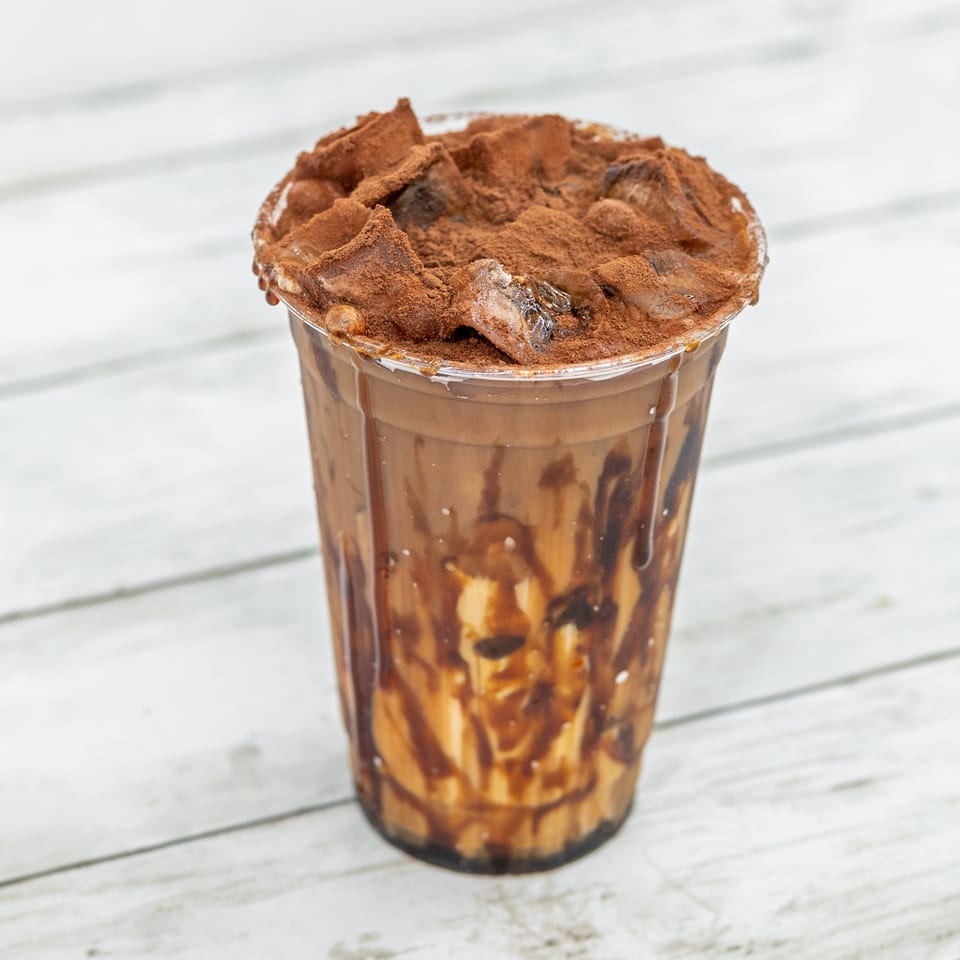 Chocolate drink in a clear cup drizzled with chocolate sauce, topped with whipped cream and cocoa powder on a white wooden table.